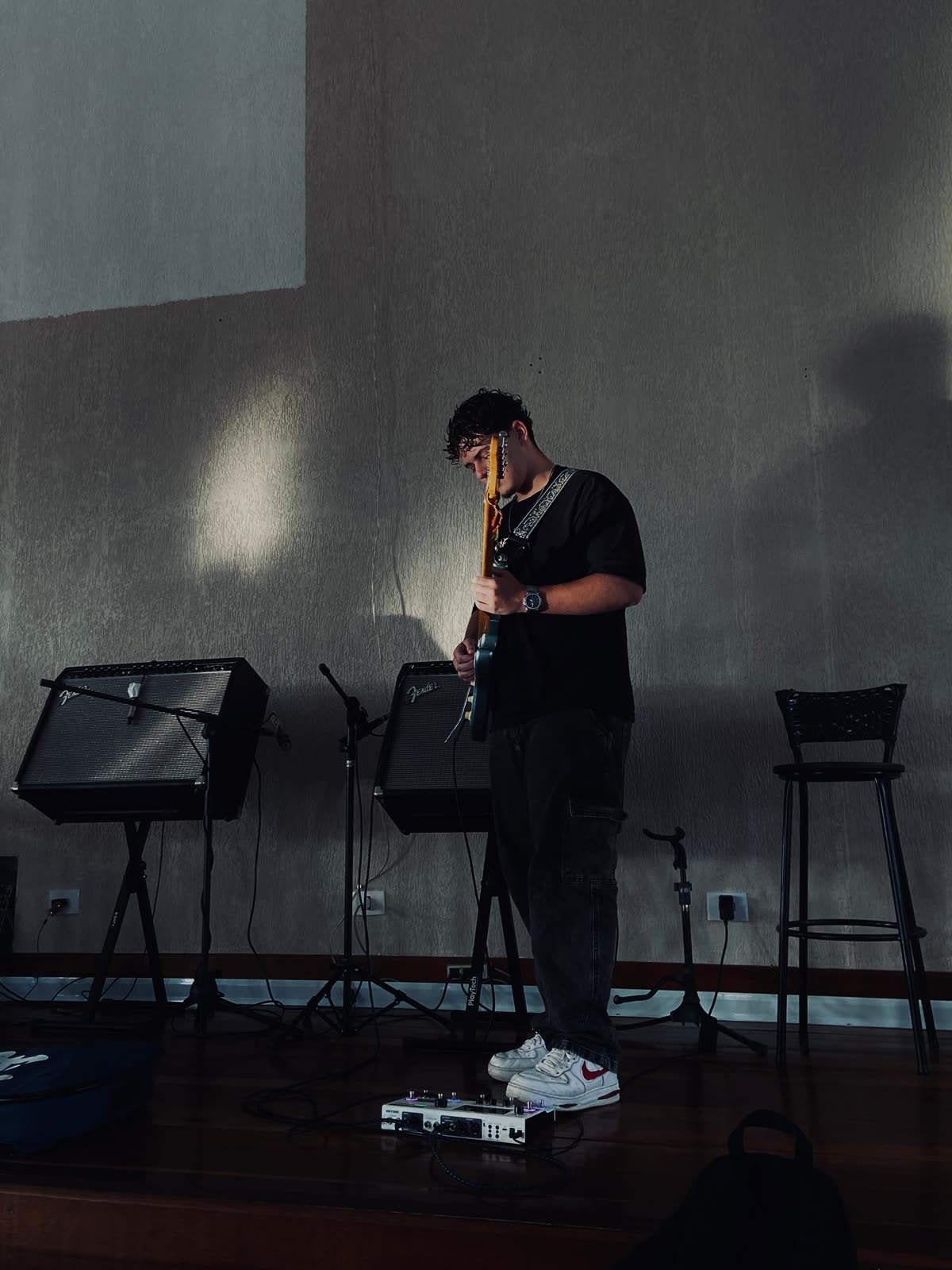 Kevin tocando guitarra em pé, numa sala com amplificadores
